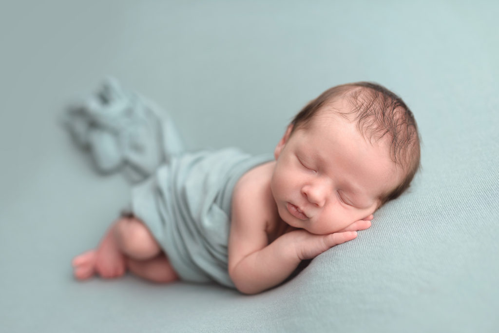 during his newborn session in Cedar Rapids, a sleeping baby boy is wrapped in a teal blanket 
