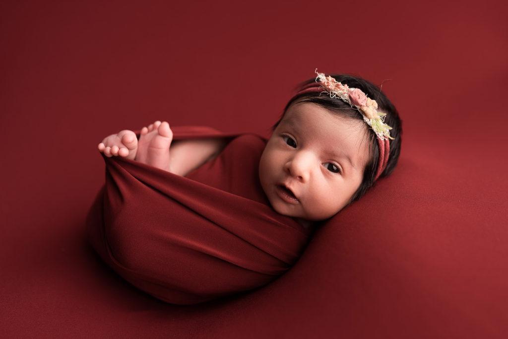 A newborn baby girl is awake on a red blanket during her photo session with Rebekah Stewart Photography in Cedar Rapids