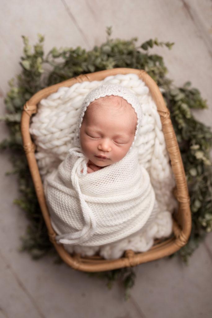 a newborn baby girl wrapped in white sleeps in a basket with greenery during her photographer session in cedar rapids
