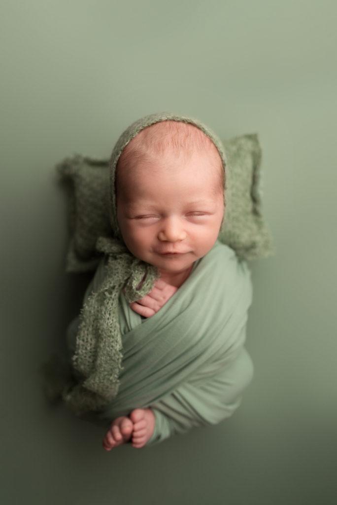 A newborn baby girl wrapped in green smiles during her photo session with Rebekah Stewart Photography in Cedar Rapids Iowa newborn must haves