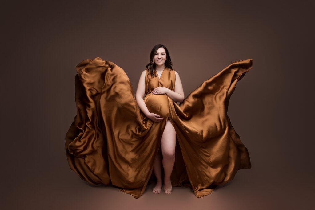 An expecting mom in a gold gown with a flying train smiles during her maternity photographer session in Cedar Rapids, maternity wardrobe